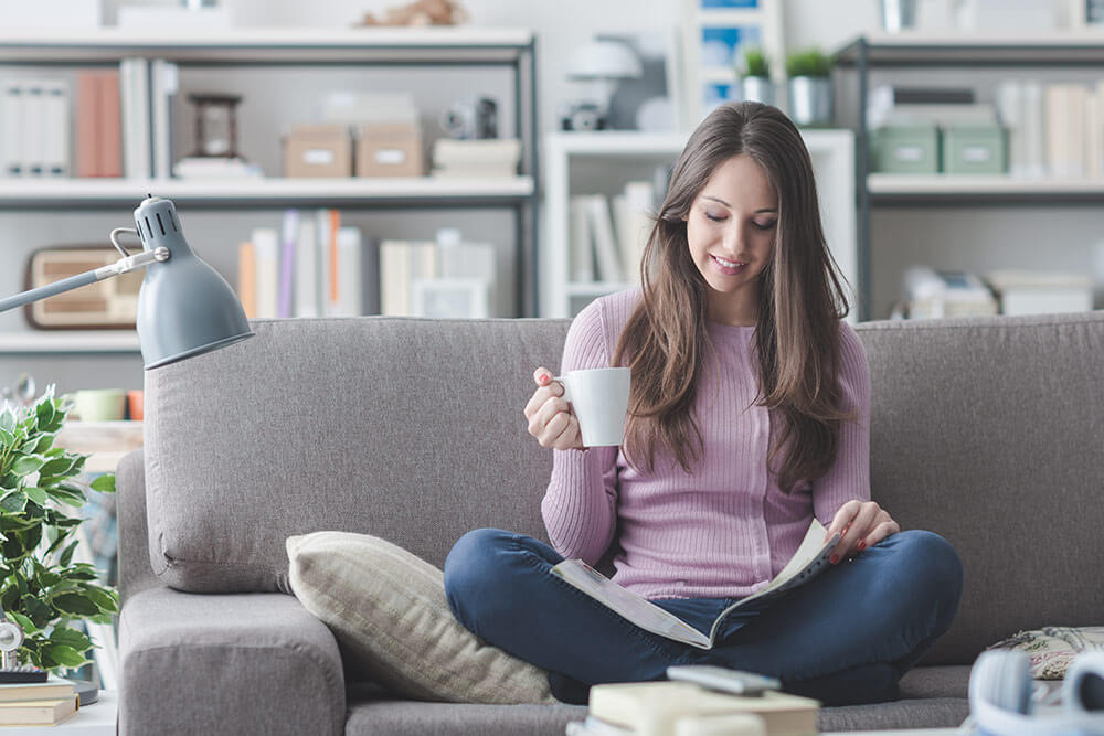 Woman with cup of coffee in hand, sitting on living room couch reading a book